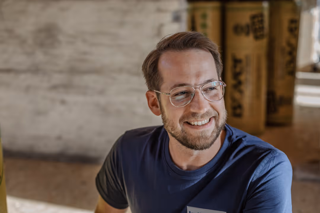 Ein lächelnder Mann mit Brille und Bart trägt ein blaues T-Shirt in einem hellen Raum.