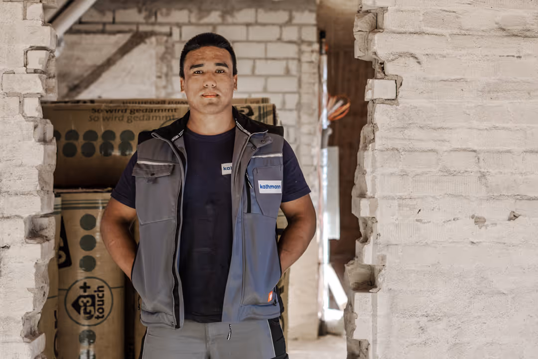Handwerker in grauer Arbeitsweste steht vor einer teilweise durchbrochenen weißen Backsteinwand auf einer Baustelle.