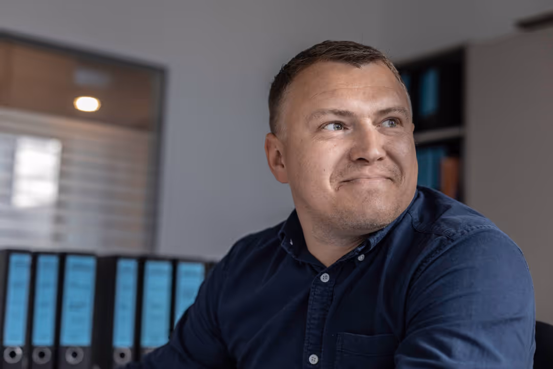 Mann in blauem Hemd sitzt vor einem Regal mit blauen Aktenordnern und schaut nach rechts.