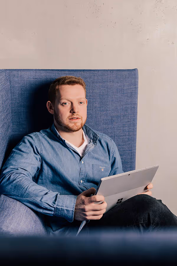 Mann im blauen Hemd sitzt auf einem blauen Sessel und hält ein Tablet.
