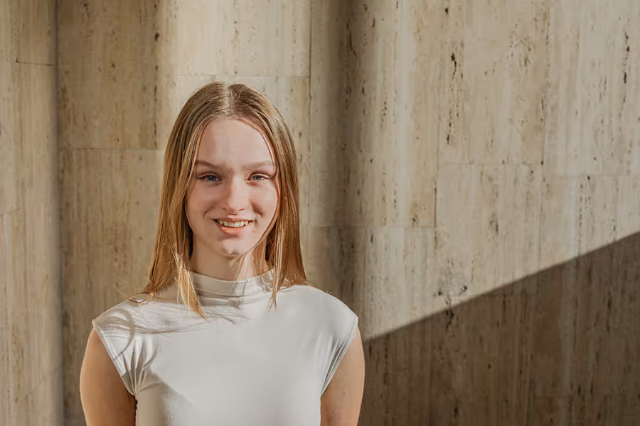 Junge Frau mit langen blonden Haaren und weißem Oberteil lächelt vor einer Holzpaneelwand.