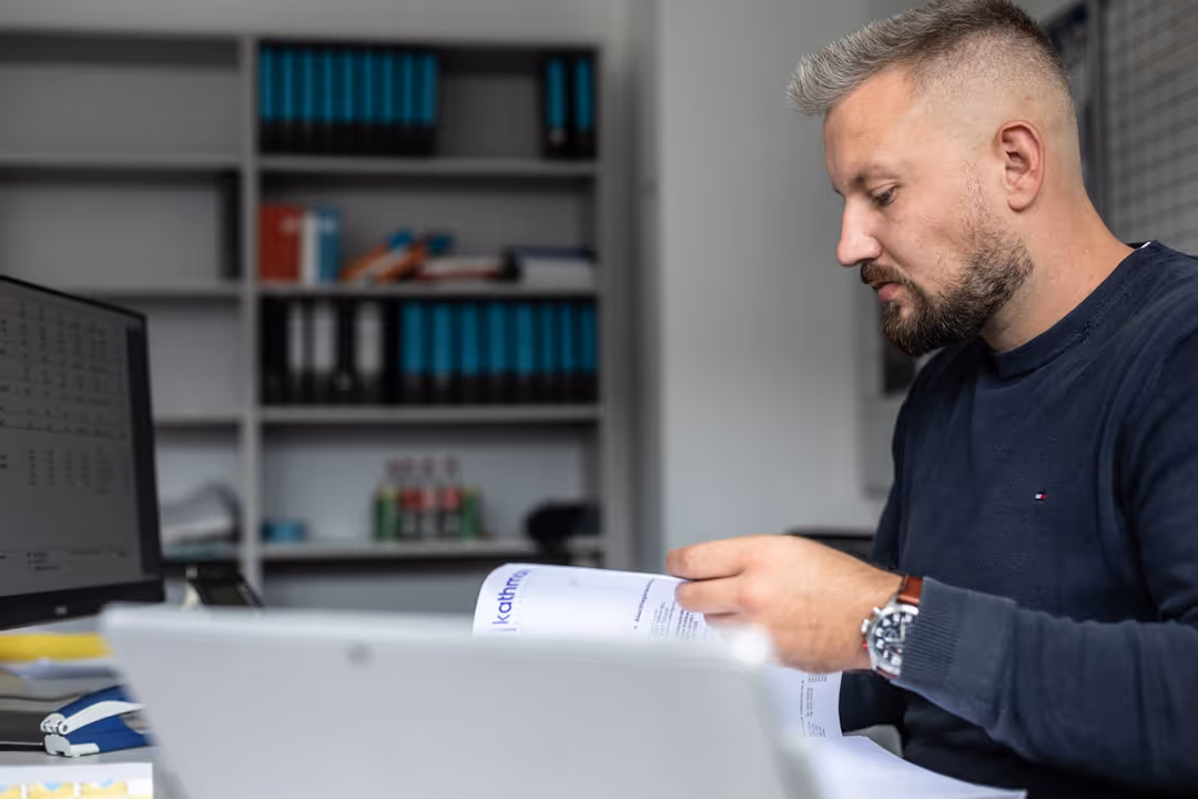 Mann mit Bart sitzt an einem Schreibtisch und liest konzentriert ein Dokument in einem hellen Büro.