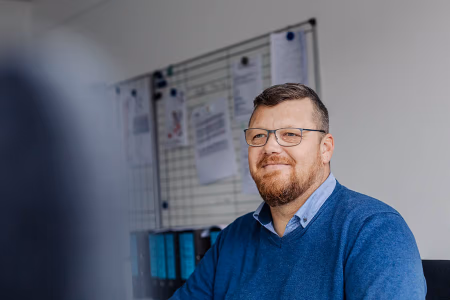 Ein Mann mit Brille und blauem Pullover lächelt in einem Büro mit weißen Wänden und einem Pinnwand-Hintergrund.