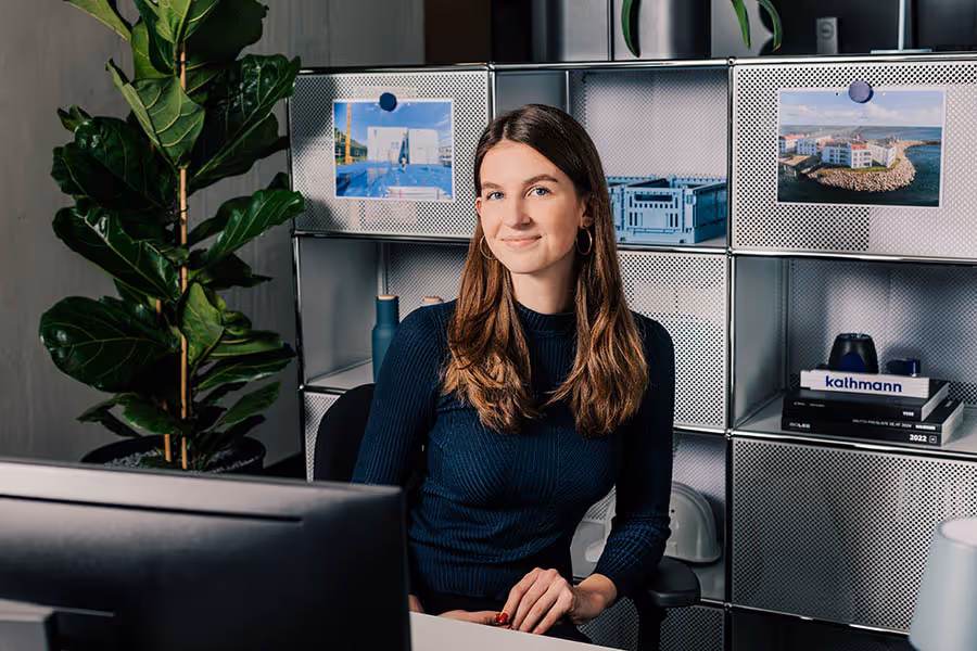 Junge Frau mit langen braunen Haaren sitzt an einem Schreibtisch vor einem Computer in einem modernen Büro mit Regalen und Pflanzen im Hintergrund.