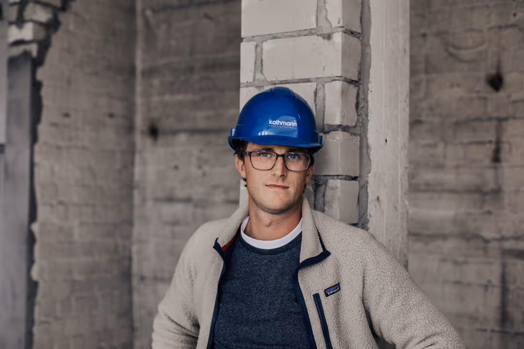 Junger Mann mit Brille und blauem Schutzhelm lehnt an einer Ziegelwand in einer Baustelle.
