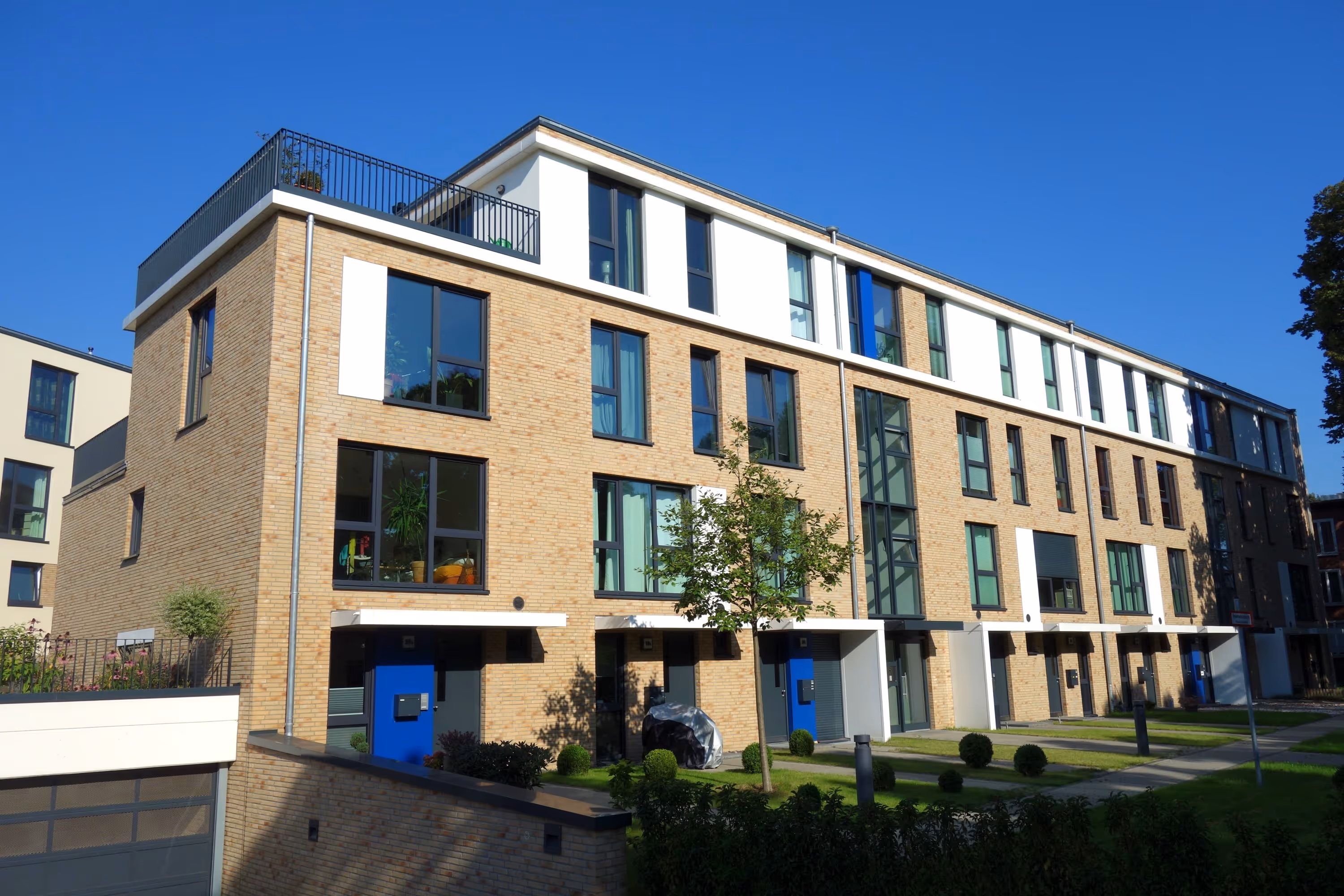 Modernes Reihenhaus mit hellen Ziegelwänden, großen Fenstern und blauen Türen unter klarem blauem Himmel.