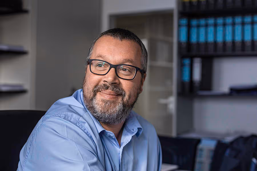 Mann mit Bart und Brille in blauem Hemd, der lächelnd zur Seite schaut, in einem Büro mit Aktenordnern im Hintergrund.