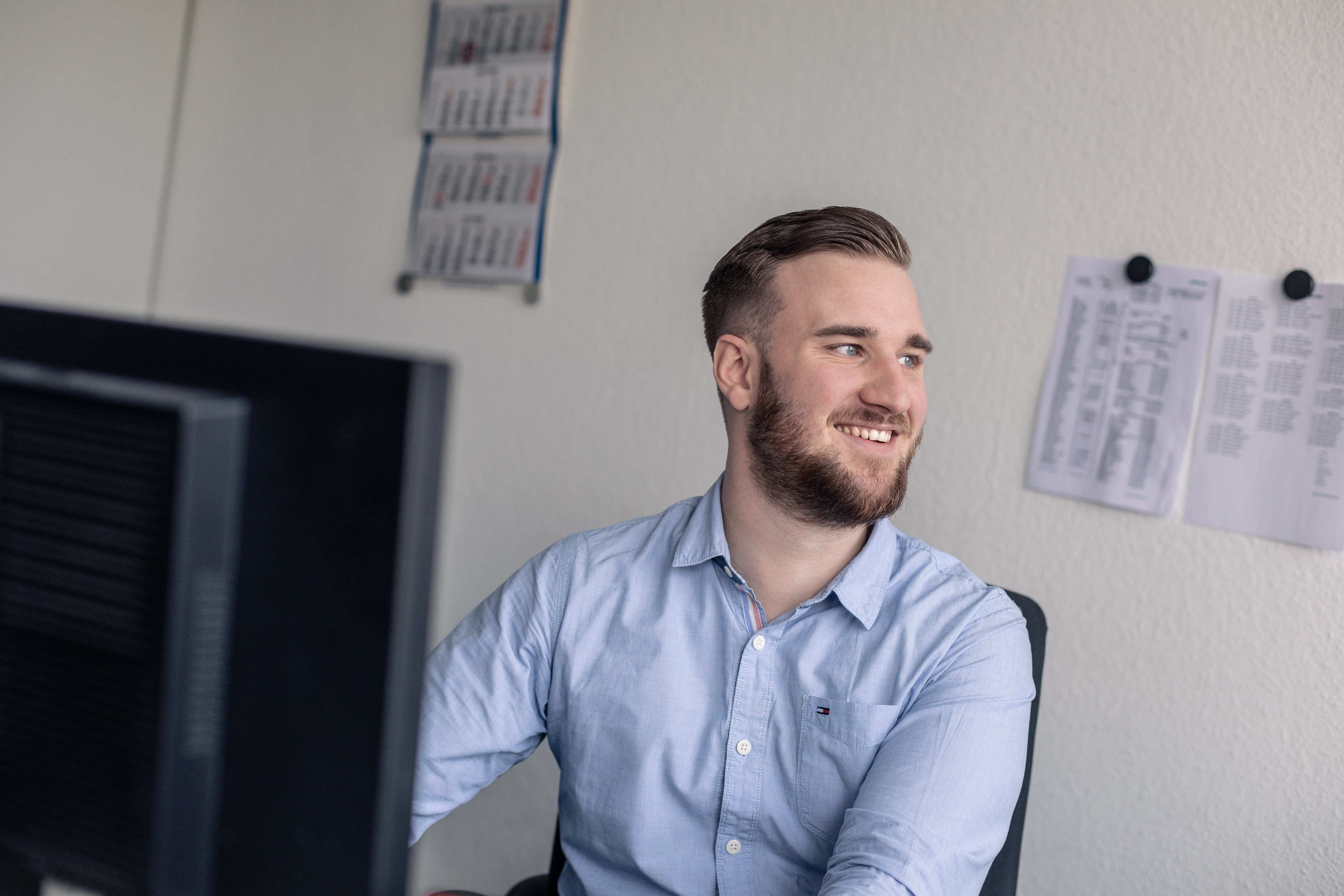Junge Frau mit langen braunen Haaren sitzt an einem Schreibtisch vor einem Computer in einem modernen Büro mit Regalen und Pflanzen im Hintergrund.
