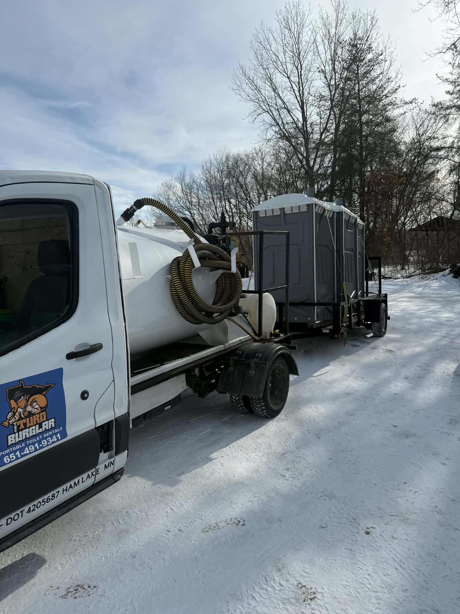 White truck with a logo for portable toilet rentals towing a trailer with two gray portable toilets on a snowy road with bare trees in the background.