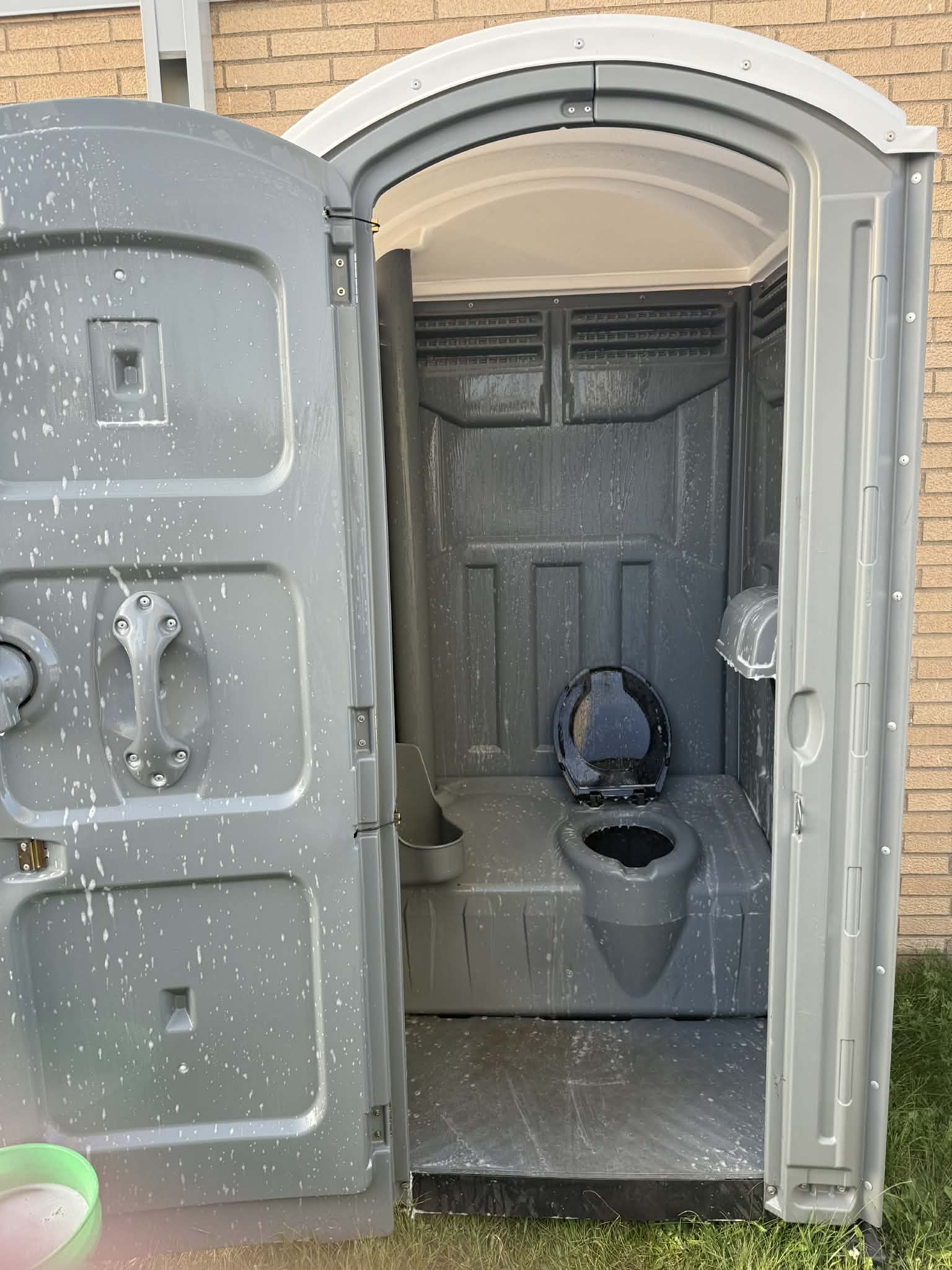 Open gray portable toilet with visible toilet seat and interior walls, situated on grass in front of a brick wall.