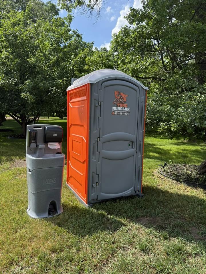 Row of six green portable toilets with white roofs lined up outdoors on an asphalt surface, with a gray portable sink unit in front and trees in the background under a blue sky.
