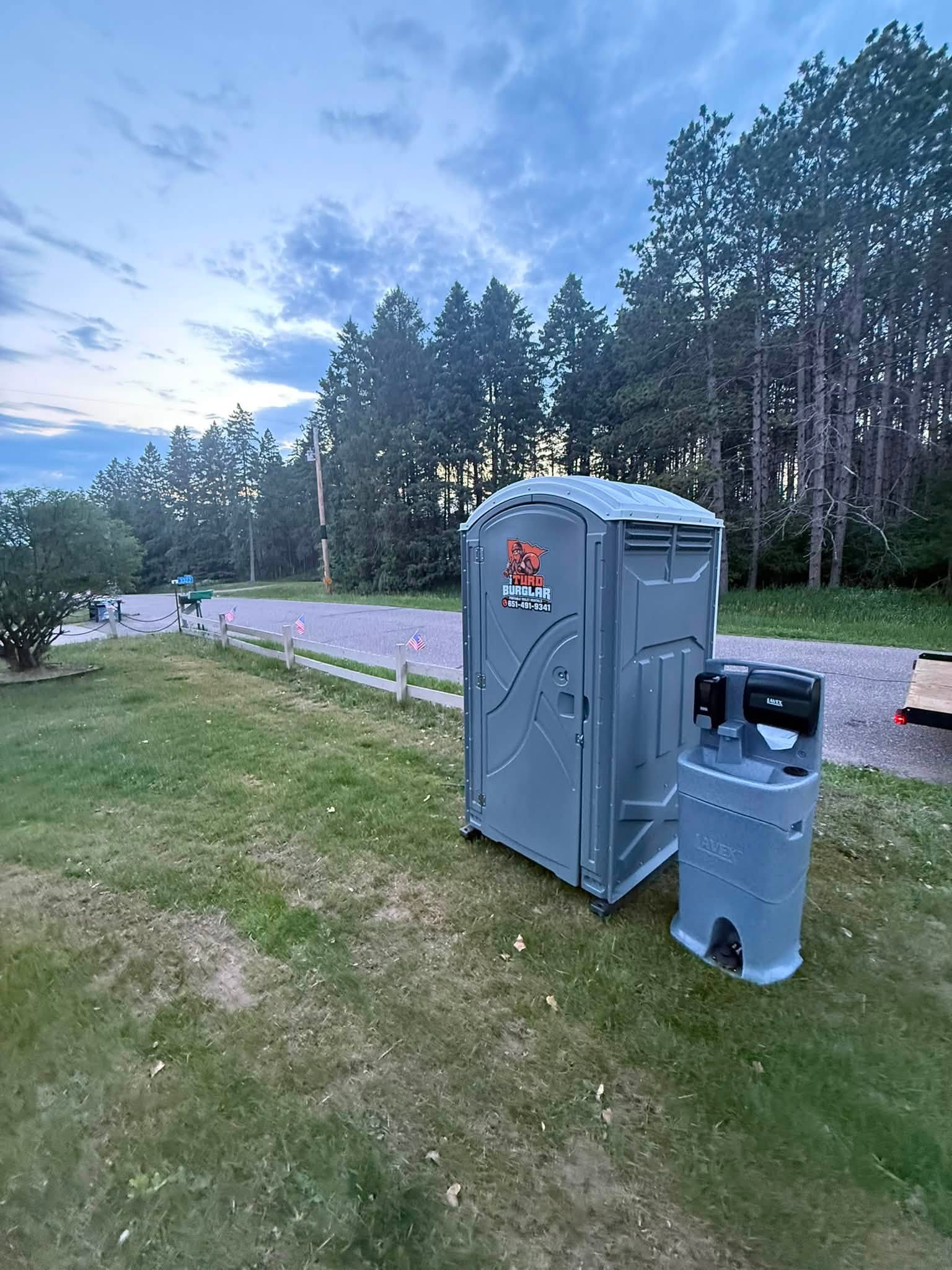 Red sanitation truck carrying green portable toilets on a grassy area under a cloudy sky.
