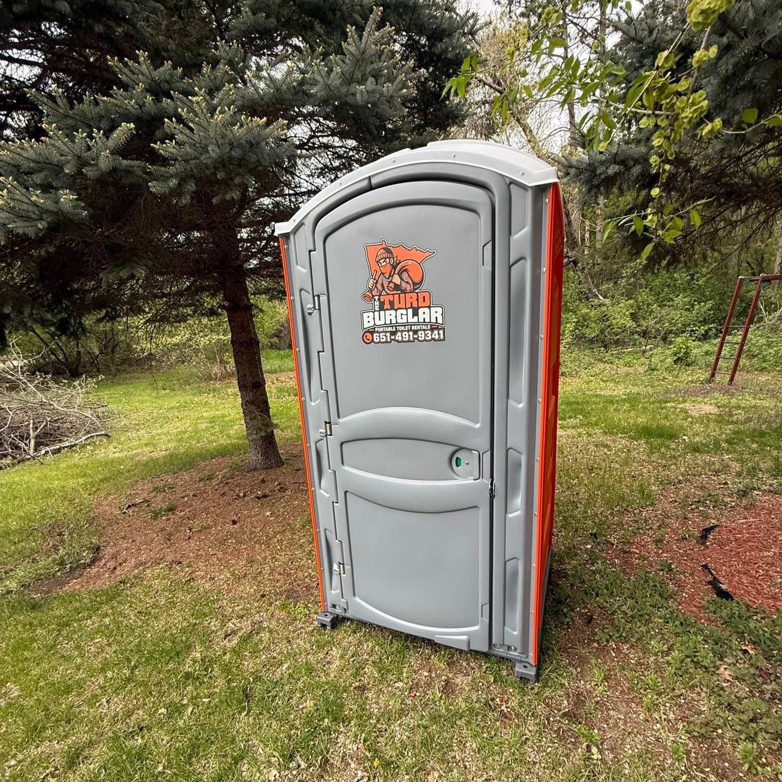 Gray portable toilet with 'Turd Burglar' logo and contact number, placed against a brick building under a cloudy sky.