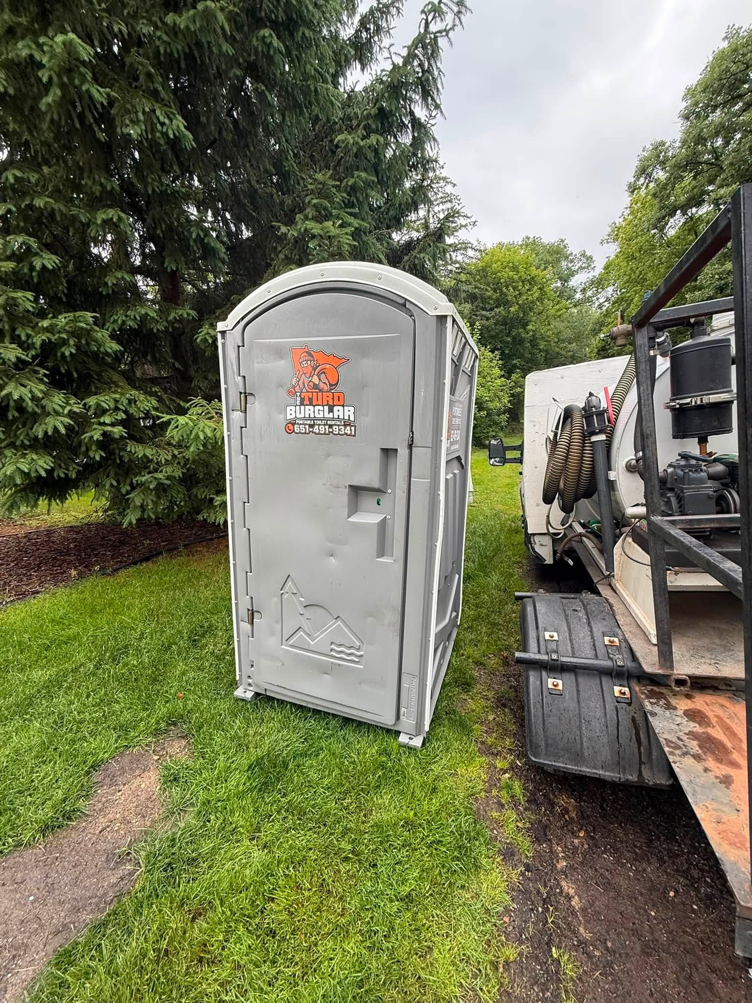 Row of green portable toilets mounted on a flatbed trailer in a grassy field under a partly cloudy sky.