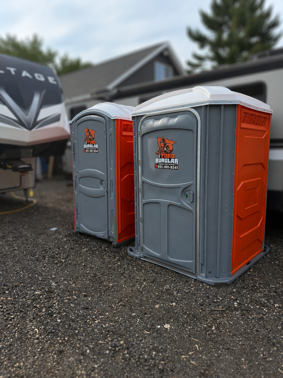 A row of seven green portable toilets with white roofs standing on wet pavement in a park-like area with trees in the background.