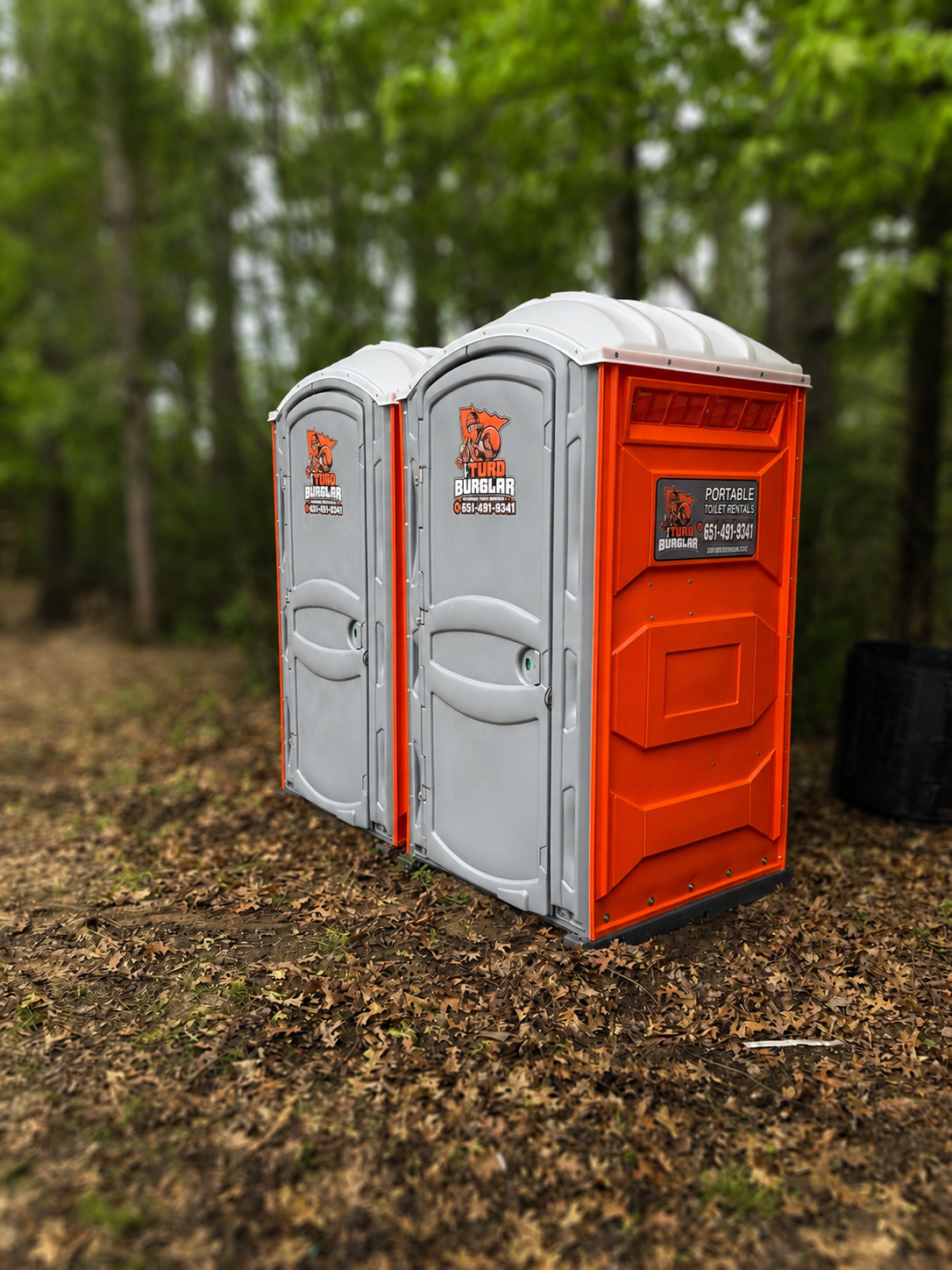 A row of seven green portable toilets with white roofs standing on wet pavement in a park-like area with trees in the background.