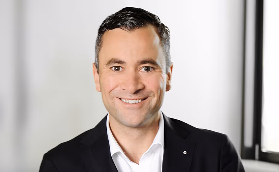 Smiling man wearing a black suit jacket and white shirt in a bright room with windows.