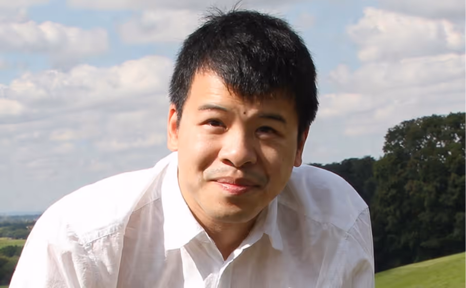 Portrait of a man in a white shirt outdoors with green fields and trees in the background.