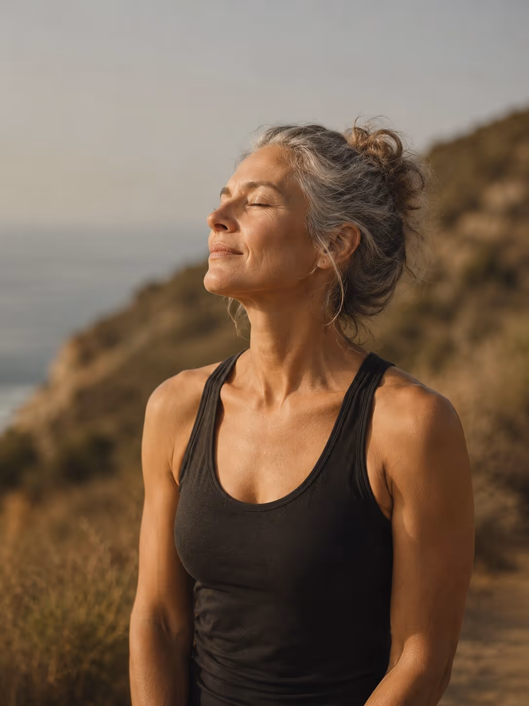 Middle-aged woman with gray hair in a bun, eyes closed, enjoying sunlight outdoors near hillside.