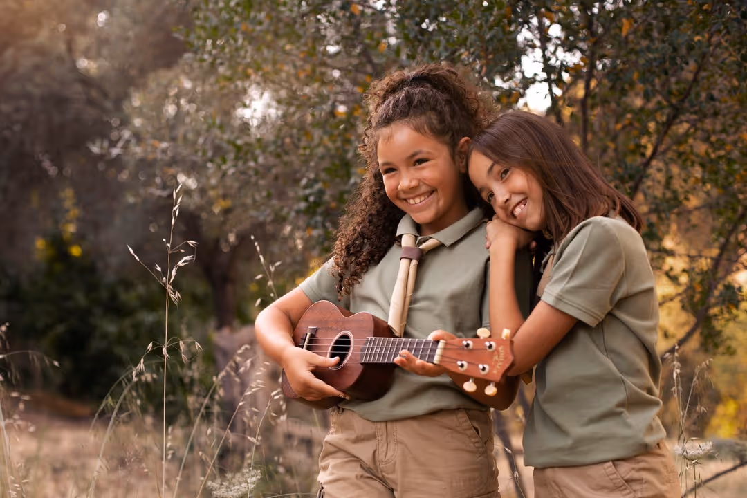 Duas meninas sorrindo ao ar livre, uma tocando ukulele e a outra apoiando a cabeça no ombro dela.