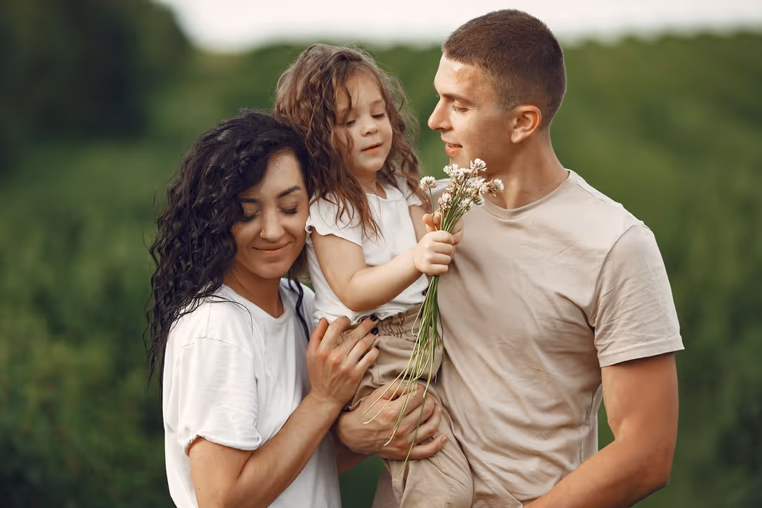 Família feliz ao ar livre, pai segurando filha com flores e mãe abraçando os dois em um campo verde.