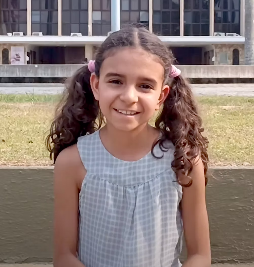 Garota sorridente com cabelos cacheados presos em dois coques e vestindo vestido xadrez cinza claro.