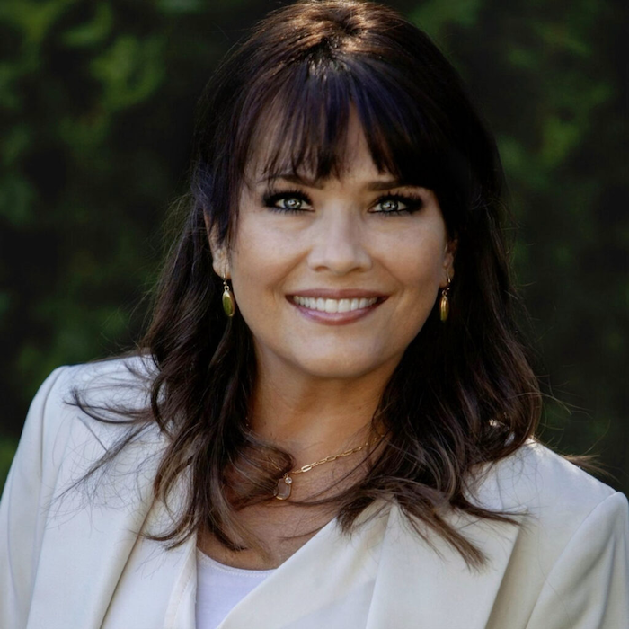 Smiling woman with dark hair, bangs, wearing gold earrings and a white blazer against a blurred green background.
