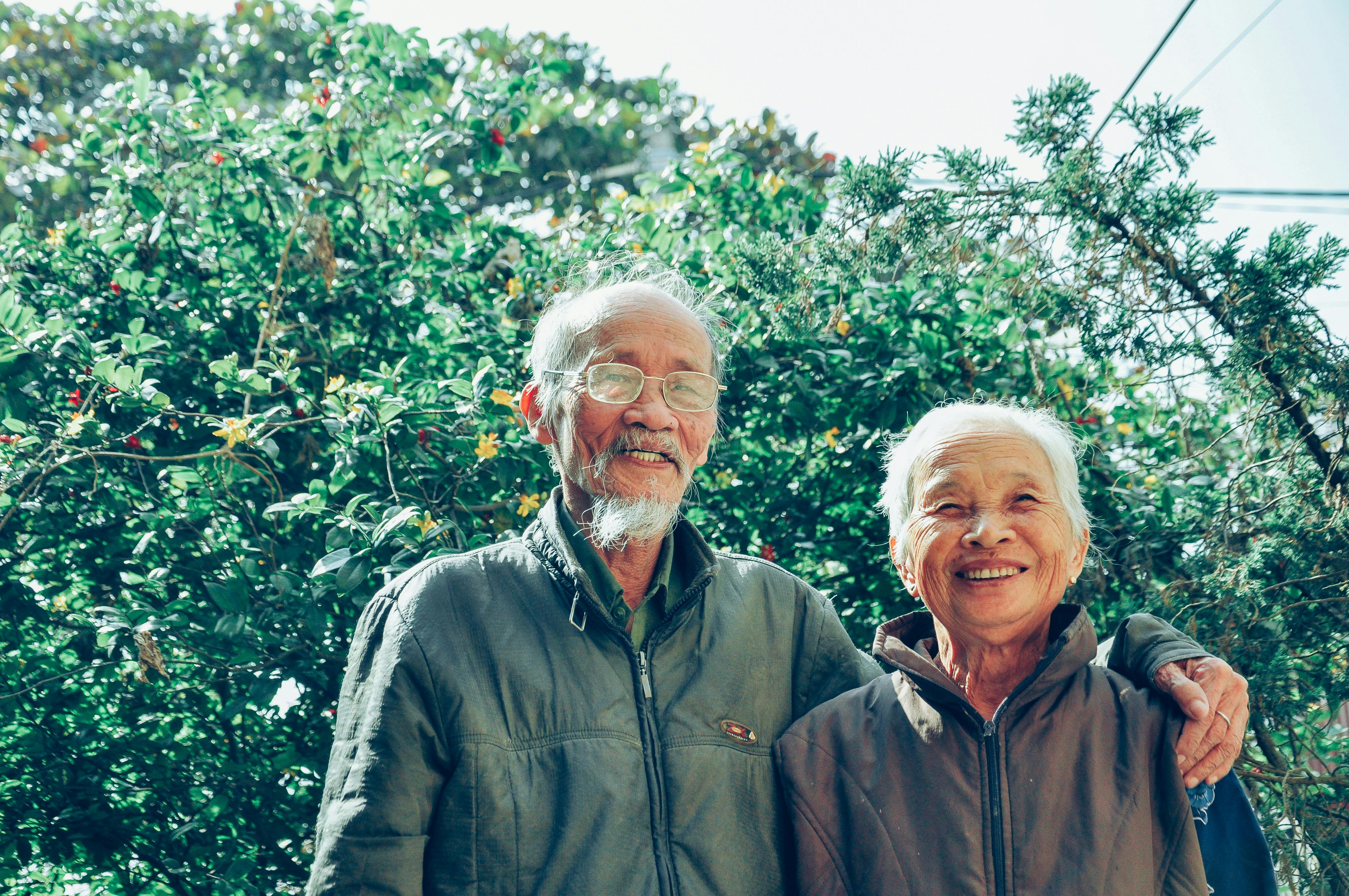 Smiling elderly couple standing outdoors in front of lush green foliage.