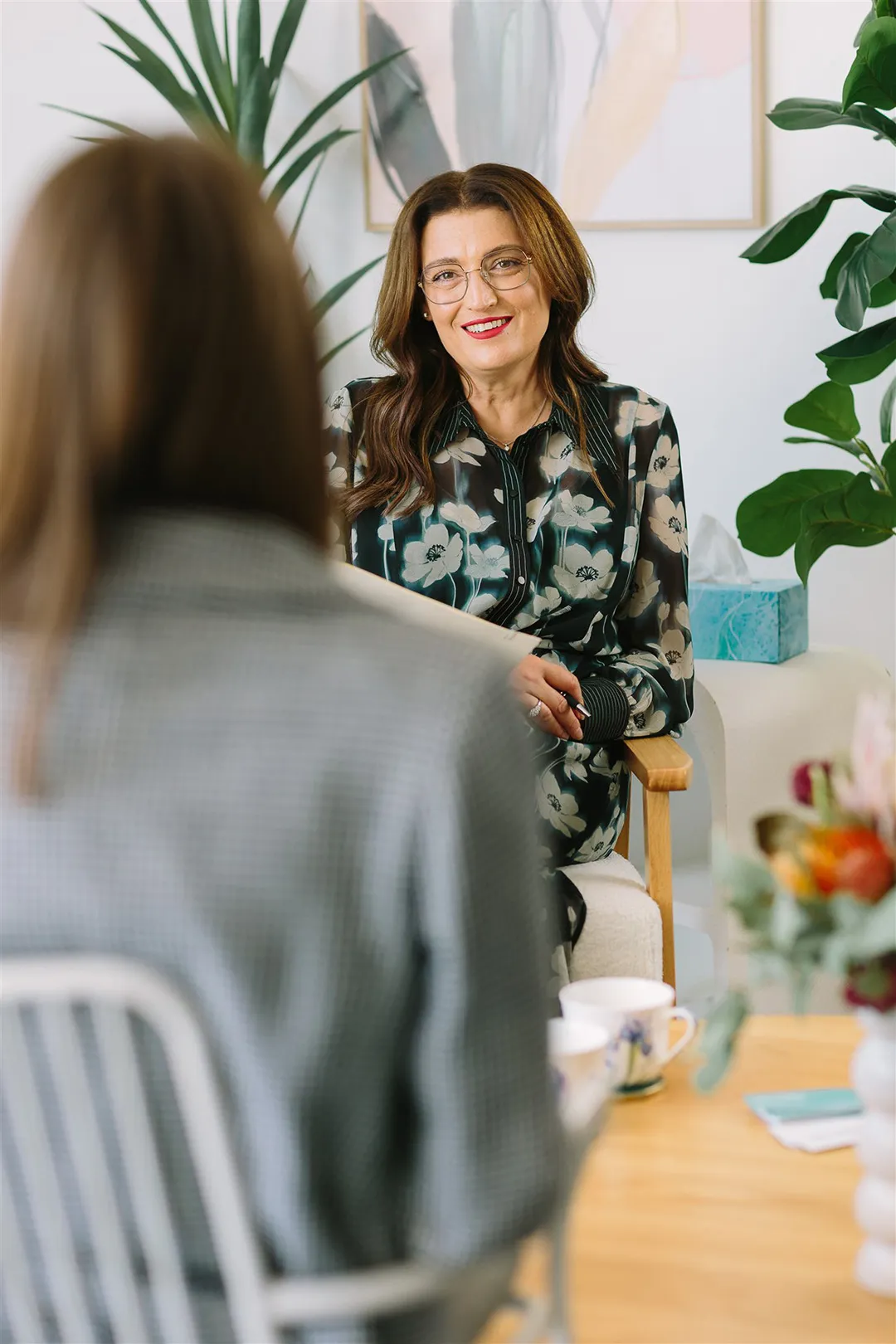Smiling woman with glasses and floral blouse sitting in a chair, speaking with a person whose back is to the camera.