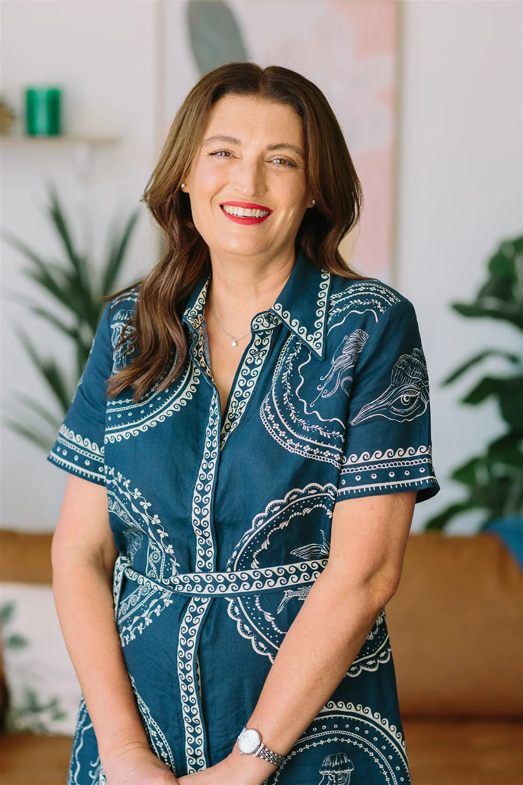 Smiling woman with brown hair wearing a blue patterned dress, standing indoors with plants and a brown sofa in the background.