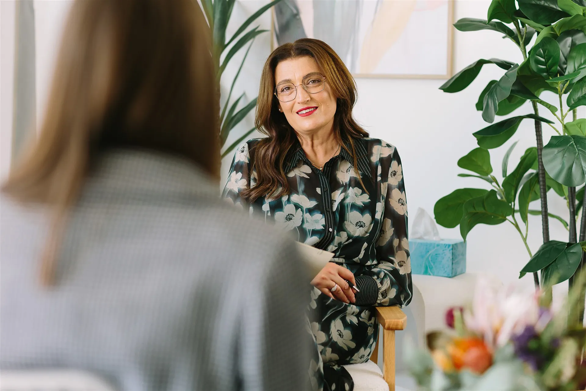 Woman with glasses and floral blouse smiling while seated and talking to another person in a cozy room with plants and flowers.