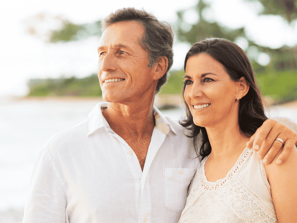 Smiling middle-aged couple standing close together outdoors with a blurred green and sandy background.