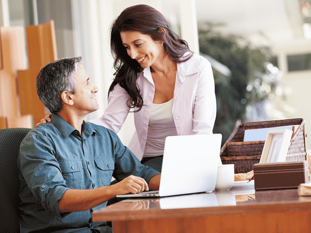 Middle-aged man sitting at a desk with a laptop looking up and smiling at a woman standing beside him with her hand on his shoulder.