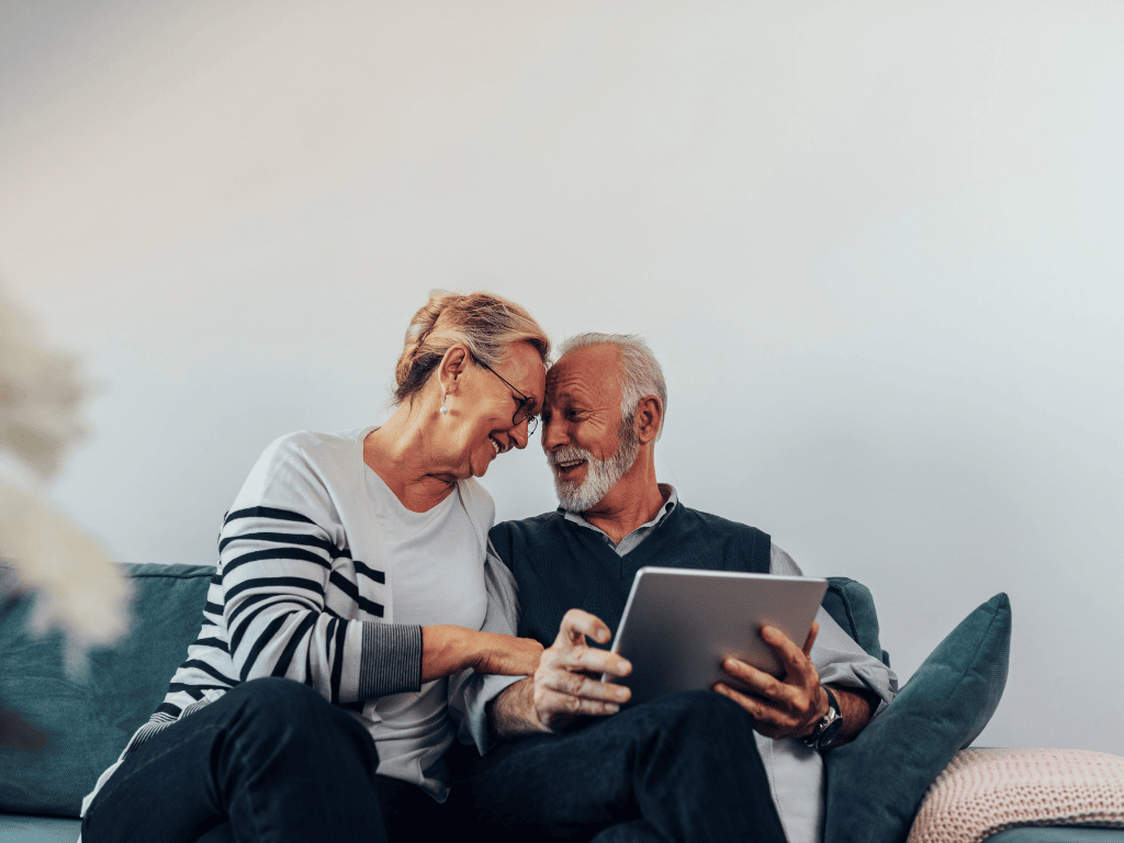 Elderly couple smiling and leaning their foreheads together while sitting on a couch with a tablet.