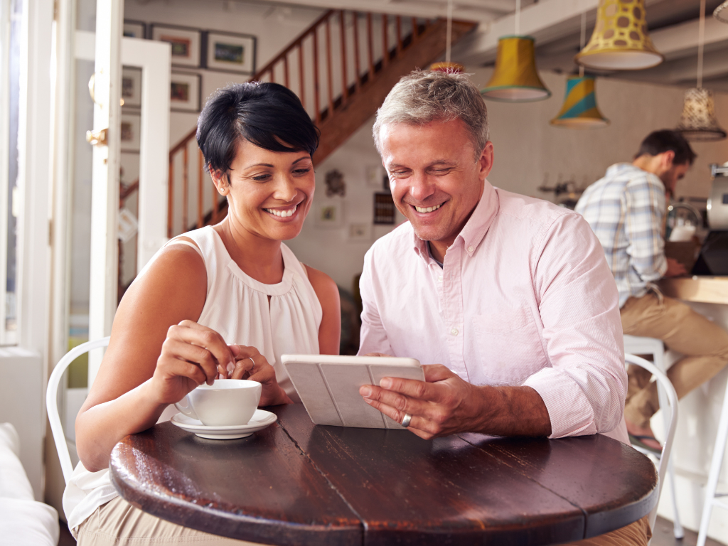 Smiling middle-aged man and woman sitting at a round wooden table in a cozy café, looking at a tablet together.