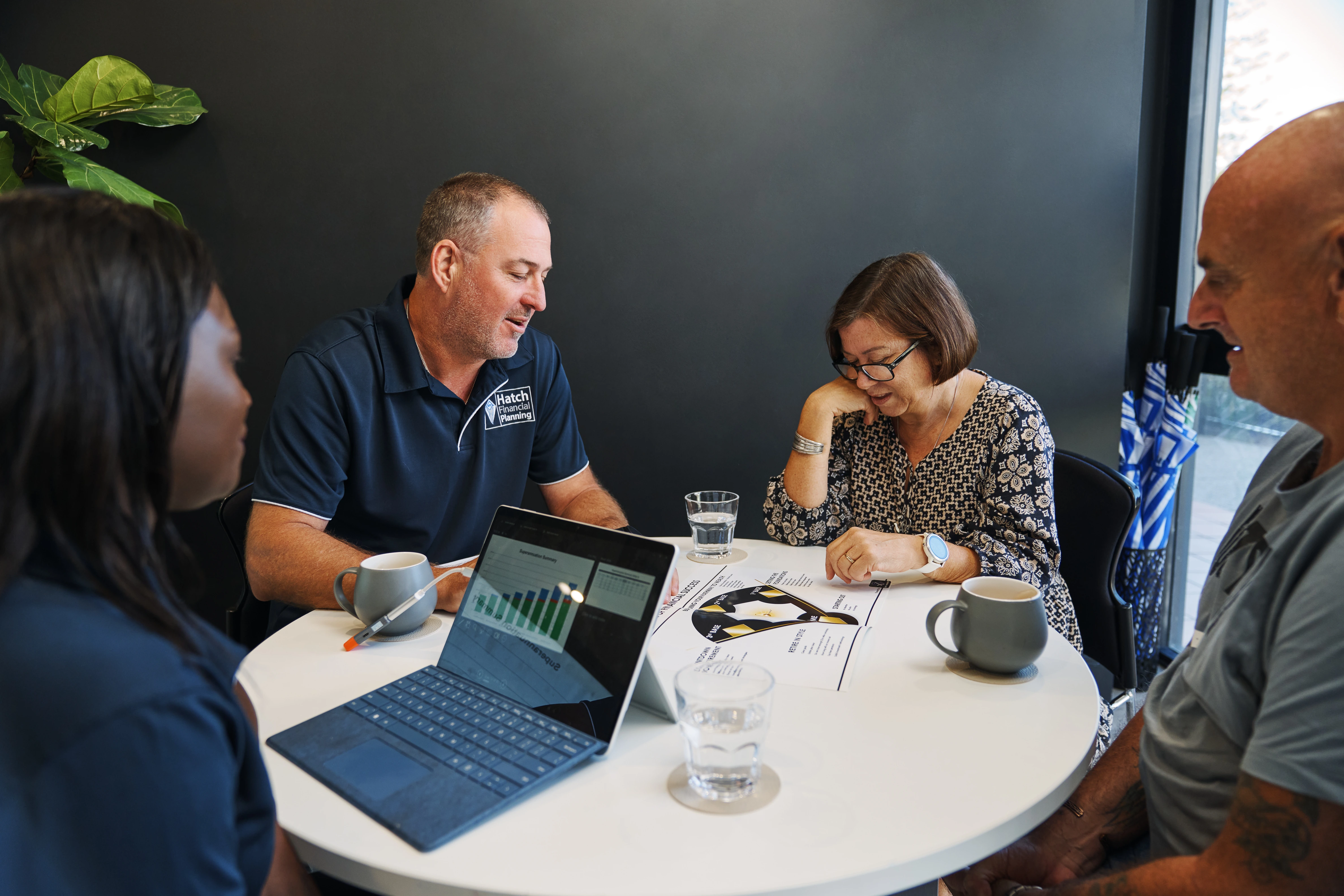 Four people in a business meeting around a round white table with charts, a laptop, coffee mugs, and glasses of water.
