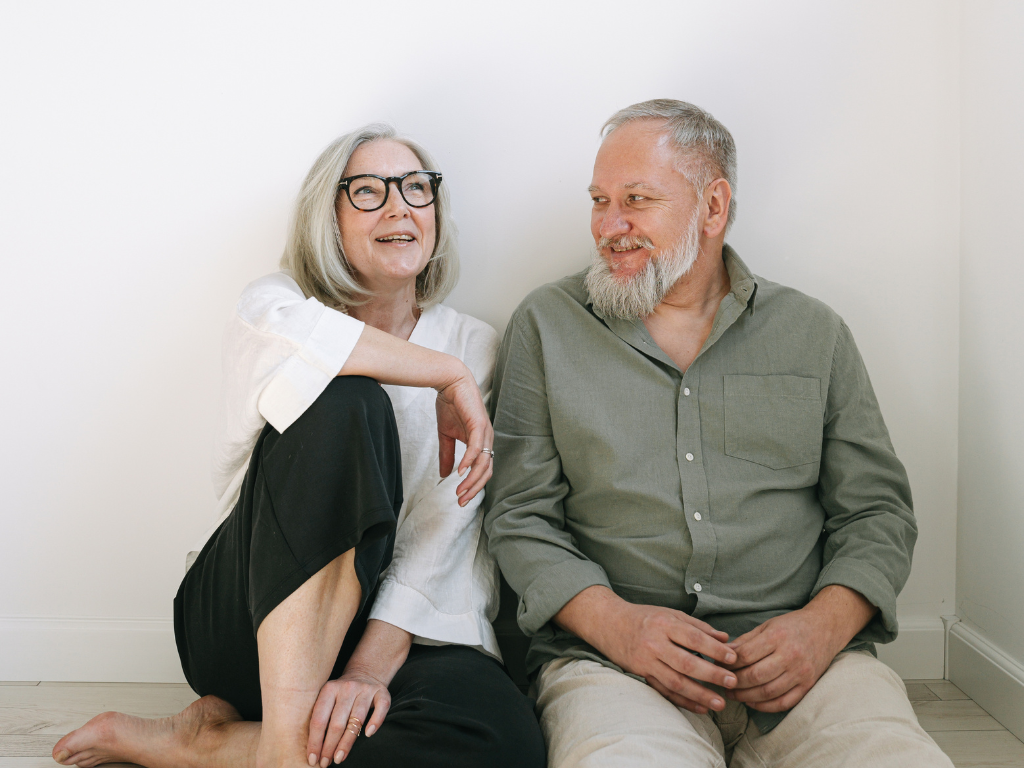 Smiling retired couple sitting on the floor against a white wall, the woman wearing glasses and a white shirt, and the man with a beard in a green shirt.