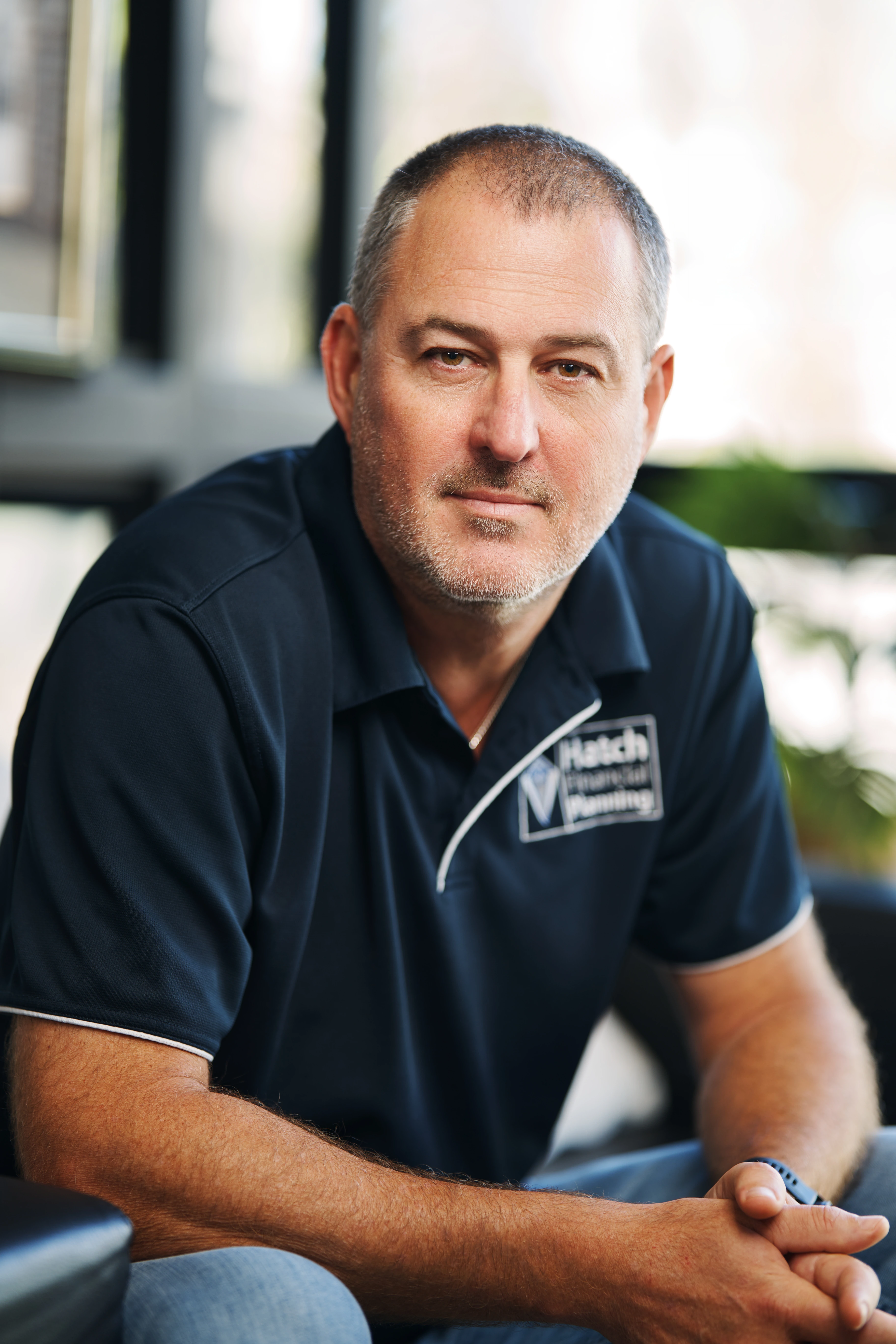 Middle-aged man with short hair and beard wearing a dark polo shirt with a visible logo sitting indoors with hands clasped.