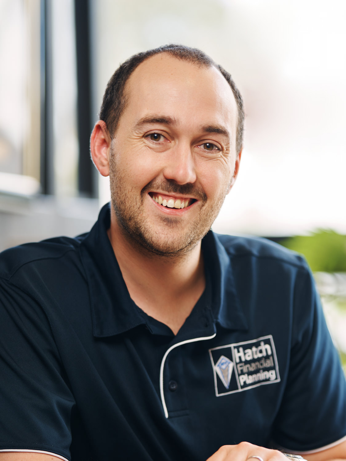 Smiling man wearing a navy blue shirt with Hatch Financial Planning logo in an office setting.