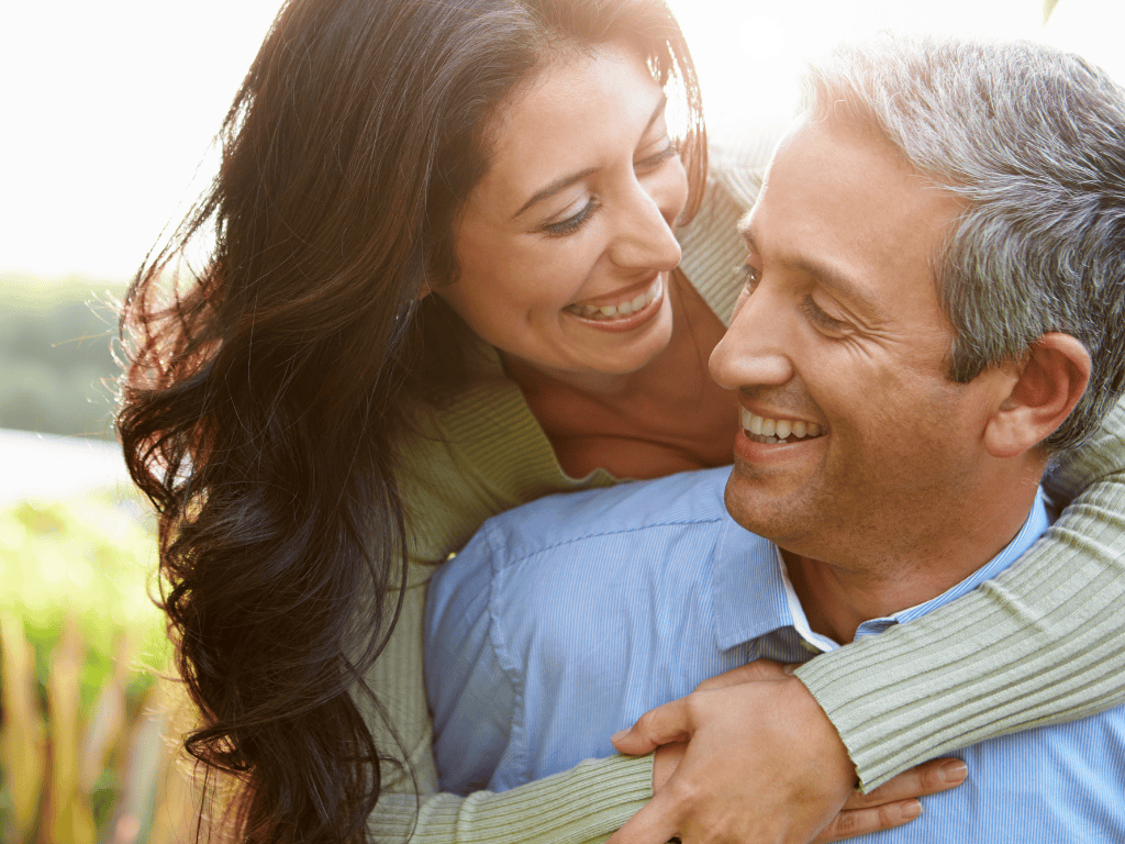 Smiling woman with long dark hair embracing a smiling gray-haired man from behind outdoors.