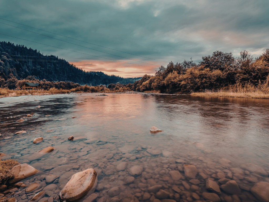 Calm river with visible rocks beneath clear water, surrounded by trees and hills under a cloudy sky at sunset.
