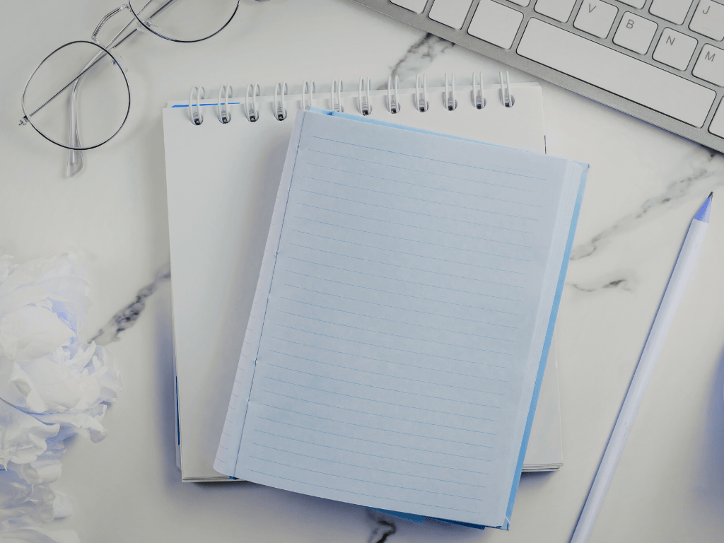 White and blue lined spiral notebooks next to round eyeglasses, a white pen with a blue tip, and part of a computer keyboard on a white marble surface.