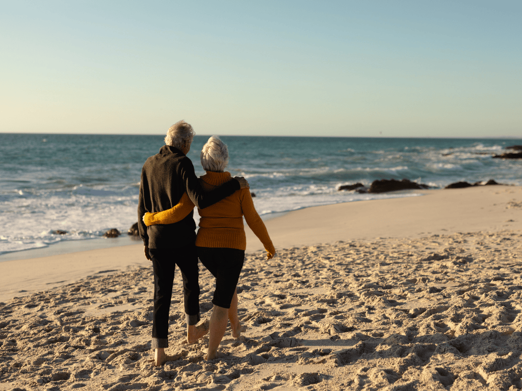 Older couple walking barefoot on a sandy beach near the ocean at sunset.