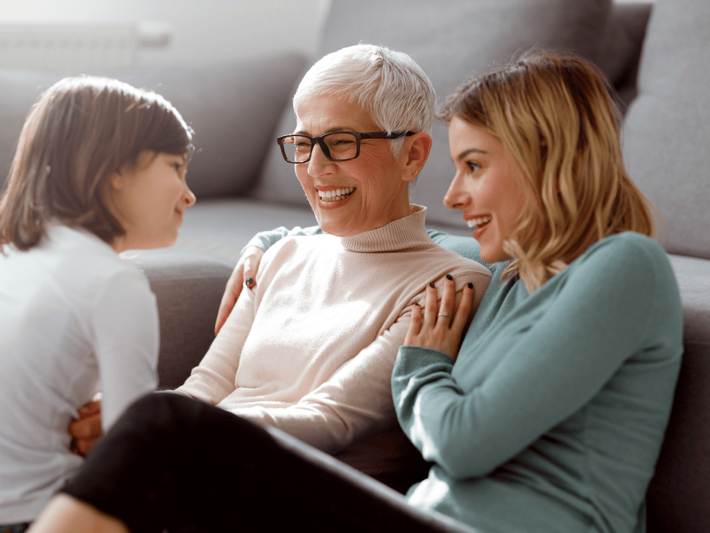 Three generations of women smiling and sharing a joyful moment indoors, sitting near a sofa.
