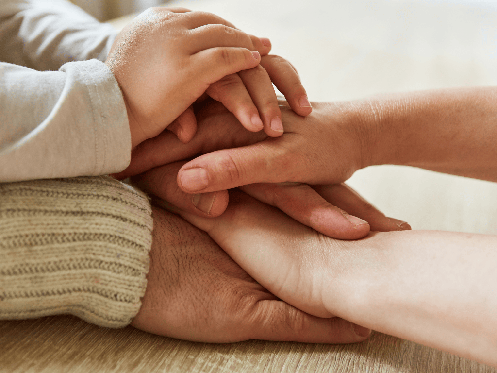 Four hands of different ages stacked together on a wooden surface symbolizing family and support.