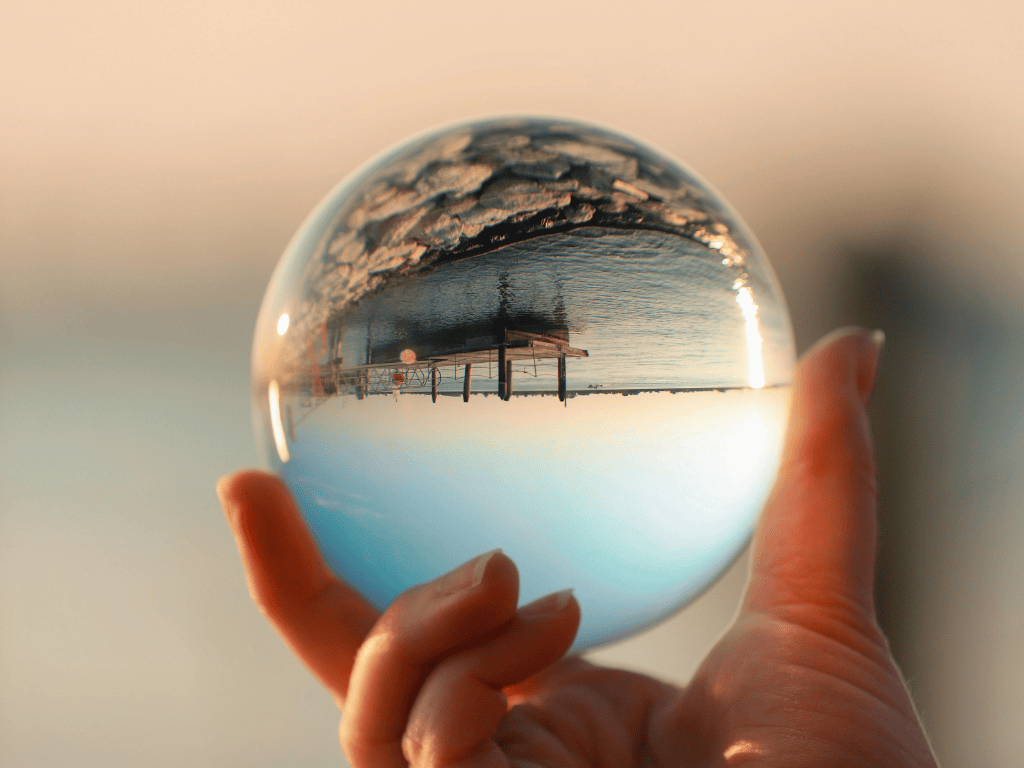 Hand holding a transparent glass sphere showing an upside-down view of a wooden pier and calm water during sunset.