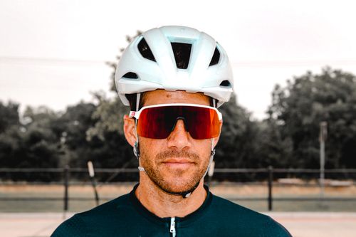 Man wearing a white cycling helmet and large red-tinted sports sunglasses, standing outdoors with blurry trees in the background.