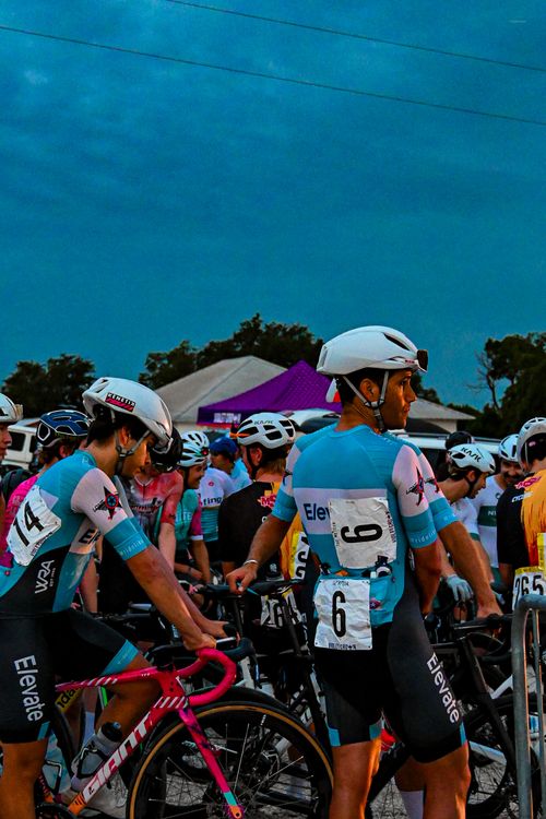 Cyclists wearing blue and black Elevate team kits with white helmets preparing at a race starting line in evening light.