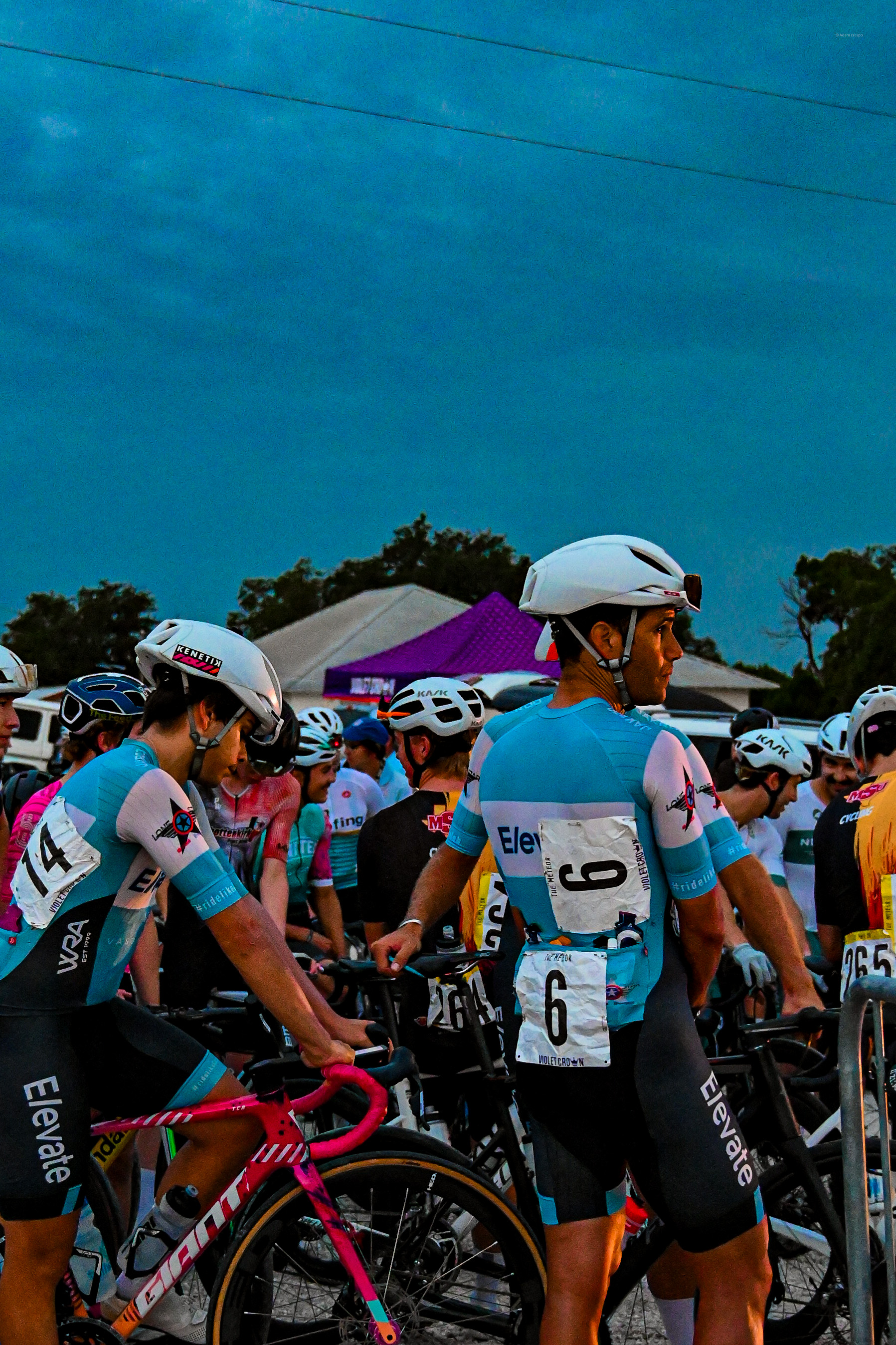 Cyclists wearing blue and black Elevate team kits with white helmets preparing at a race starting line in evening light.