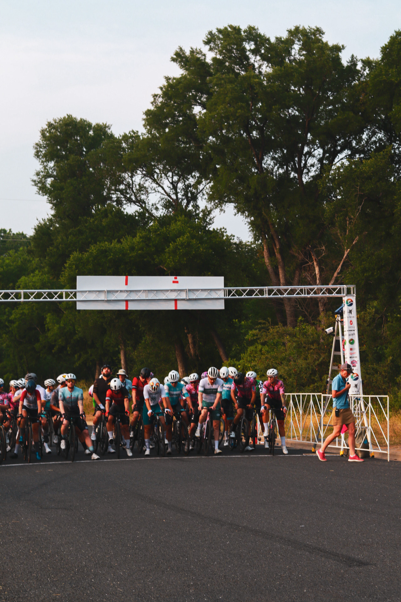 Cyclists lined up at the start line of a race on a road surrounded by trees, with a man walking nearby.